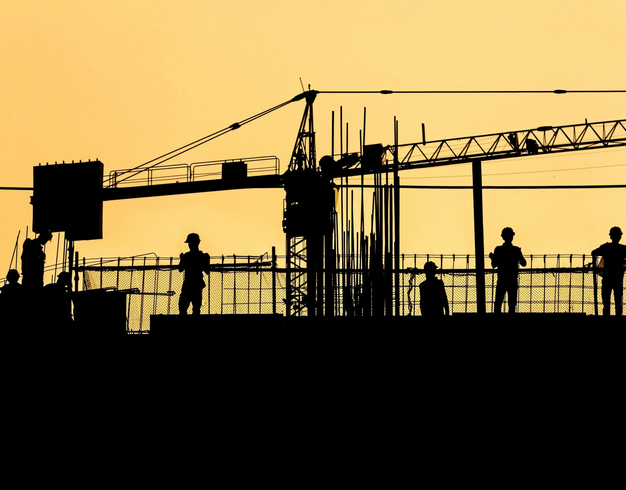 Construction workers and a tower crane silhouetted against a yellow sky at sunset, atop a partially-built structure