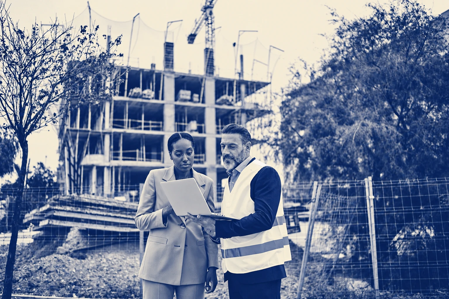 Blue-and-cream duotone photograph of two professionals reviewing a laptop beside a building under construction.