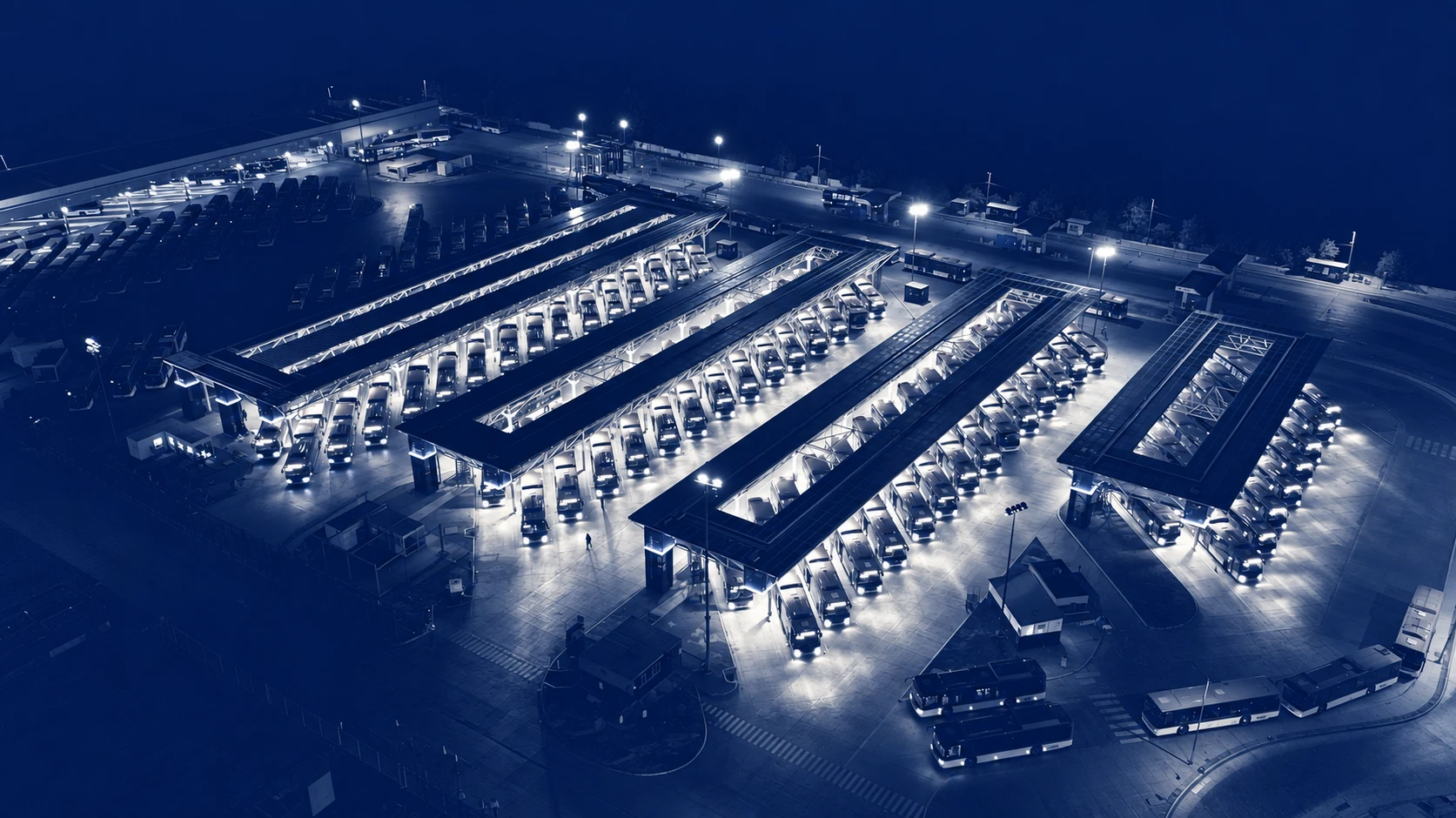 Blue-toned aerial view of a bus depot at night, with illuminated fleet rows beneath long canopies.