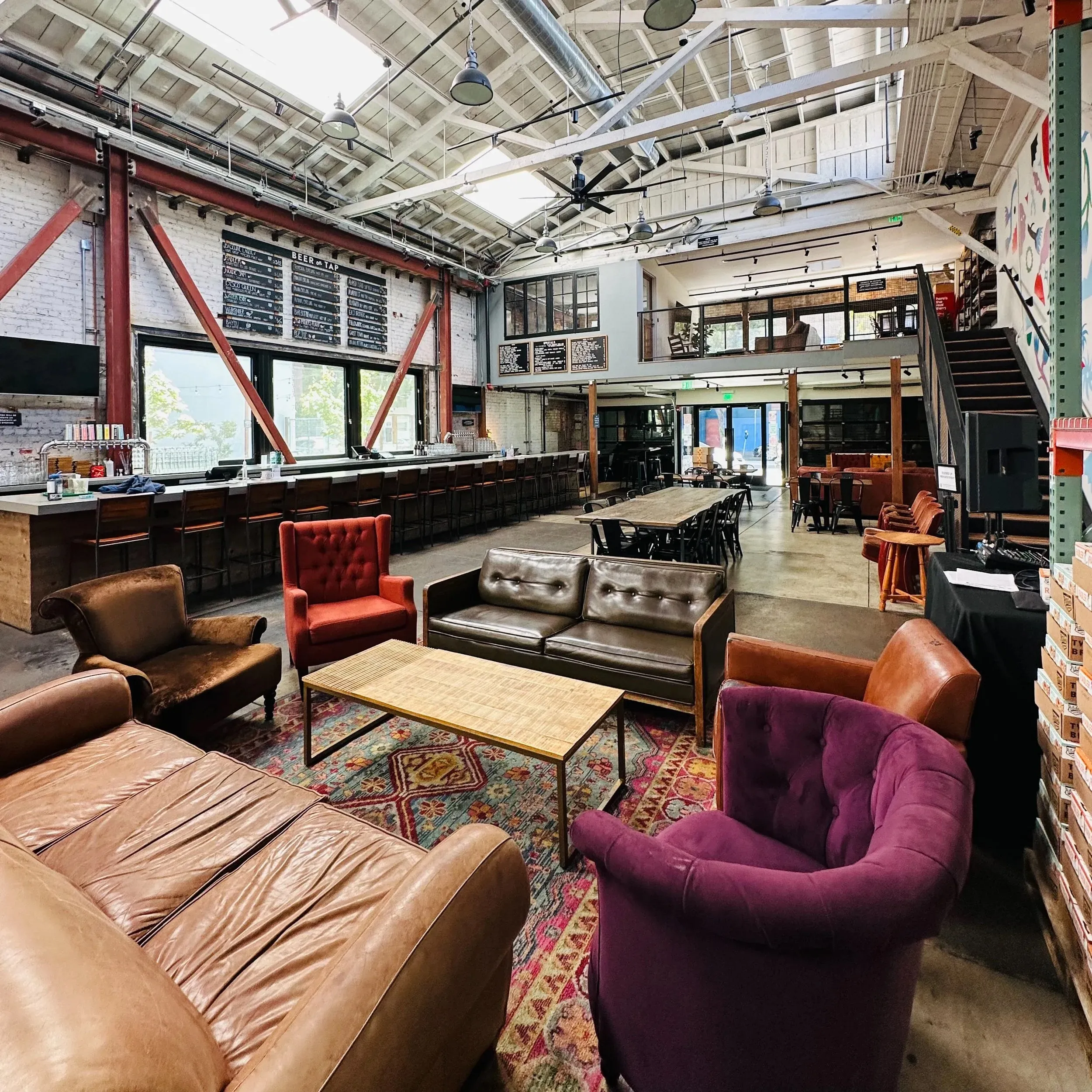 Interior of Two Pitchers' Oakland taproom, with a long bar, communal wooden tables, and leather couches beneath an exposed-brick wall and steel-truss ceiling